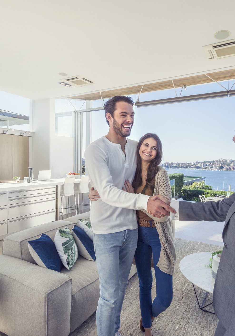 Real estate agent with couple in luxury home. They are shaking hands. There is a water view, kitchen and living room in the background. Couple are casually dressed. They are laughing. Agent is dressed in a suit and smiling. Wide angle.